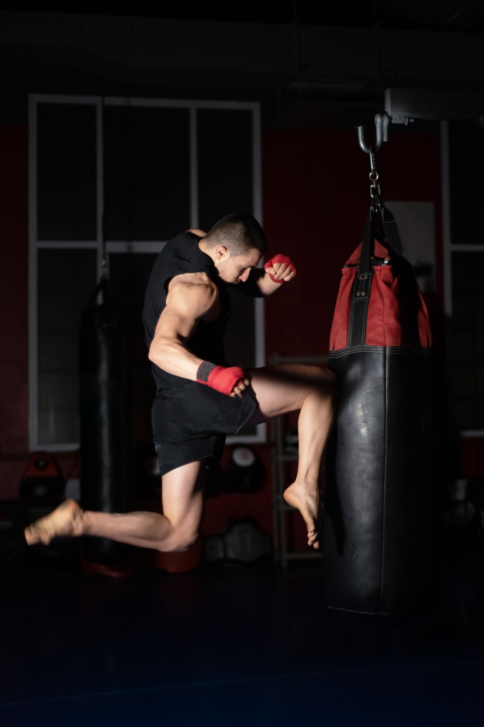 Kickbox-Kämpfer Performing Jumping Air Kicks mit Knie auf Punch Bag. Kaukasischer Mann übt Kampfkunst Training im Urban Gym.